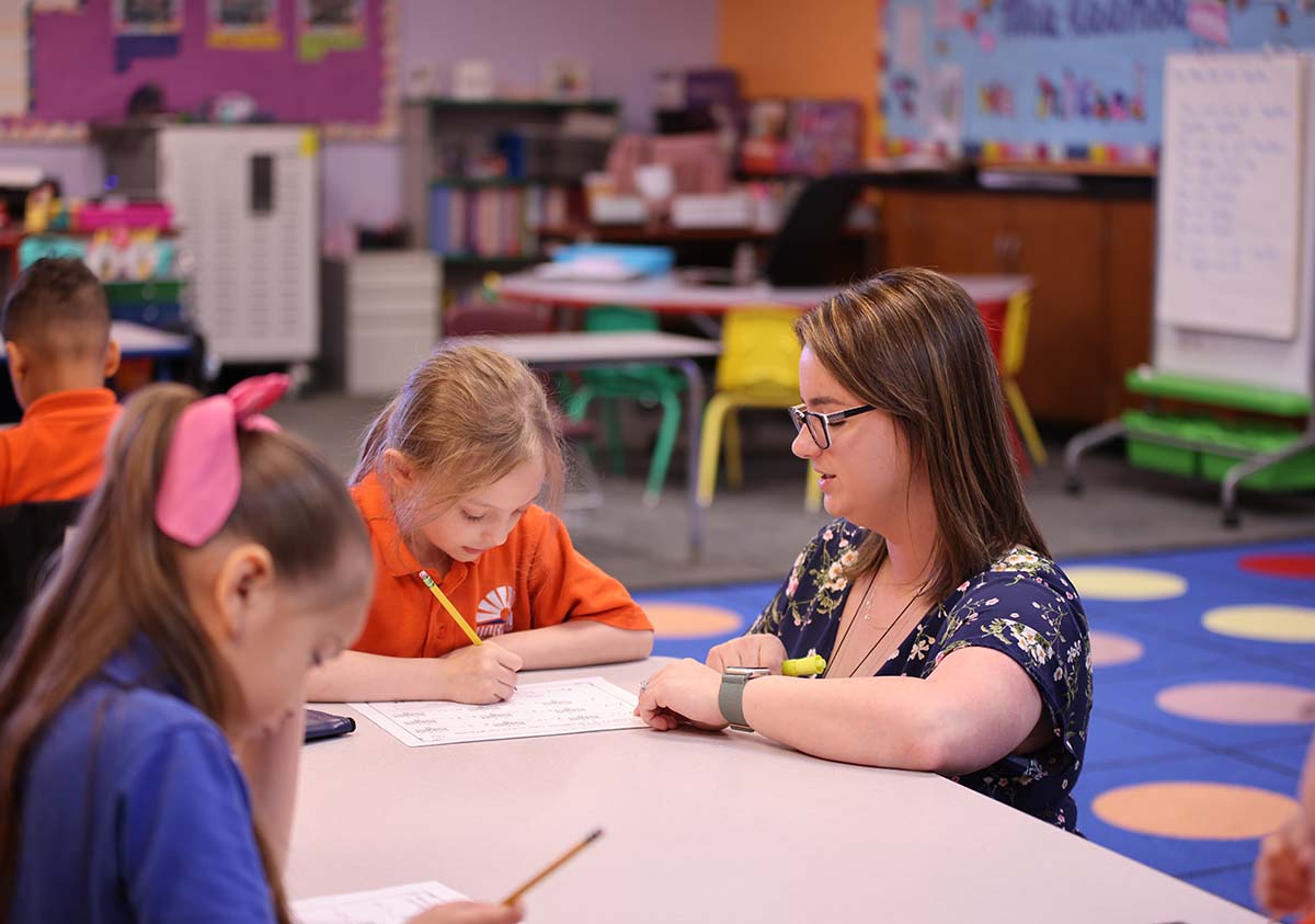 HSA Teacher smiles while kneeling beside a young student in a classroom setting.